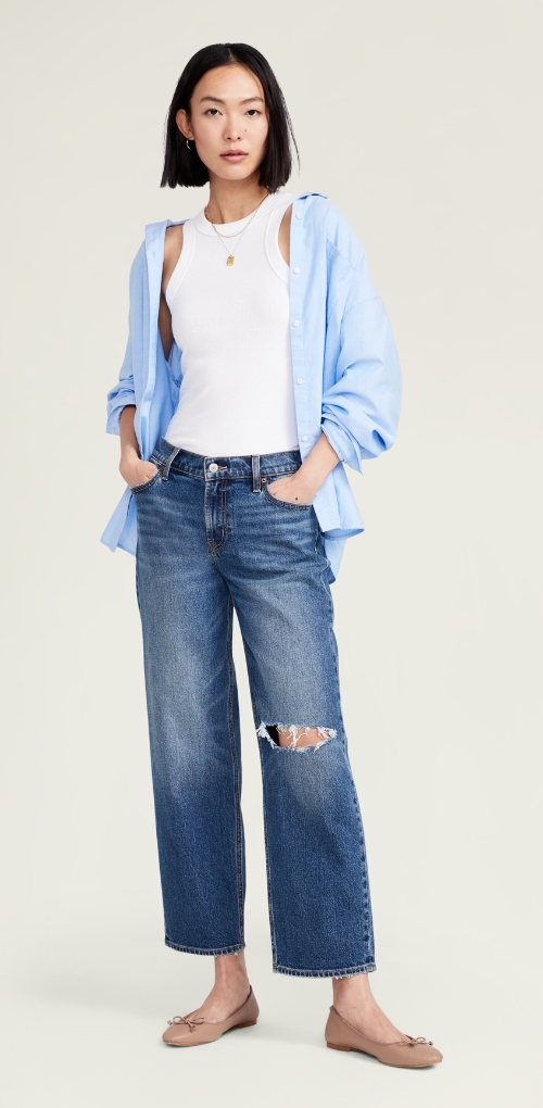 A female model in a white tank, oversized blue blouse, and loose fit boyfriend jeans with a ripped knee.