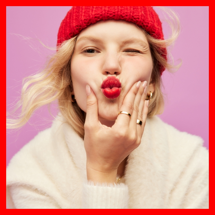 A female model blowing a kiss at the camera wearing a red beanie & cream colored sweater