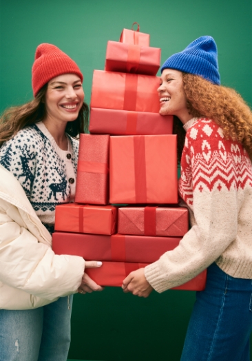 Two models wearing fair isle cardigan sweater, beanies, jeans and holding lots of presents.