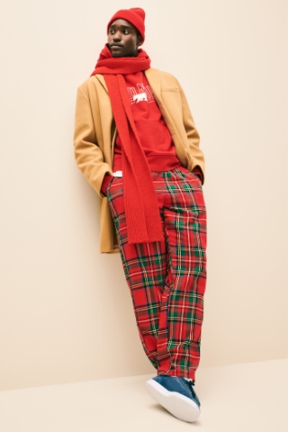 A male model wearing a holiday pajama set with a tan coat, red scarf & beanie
