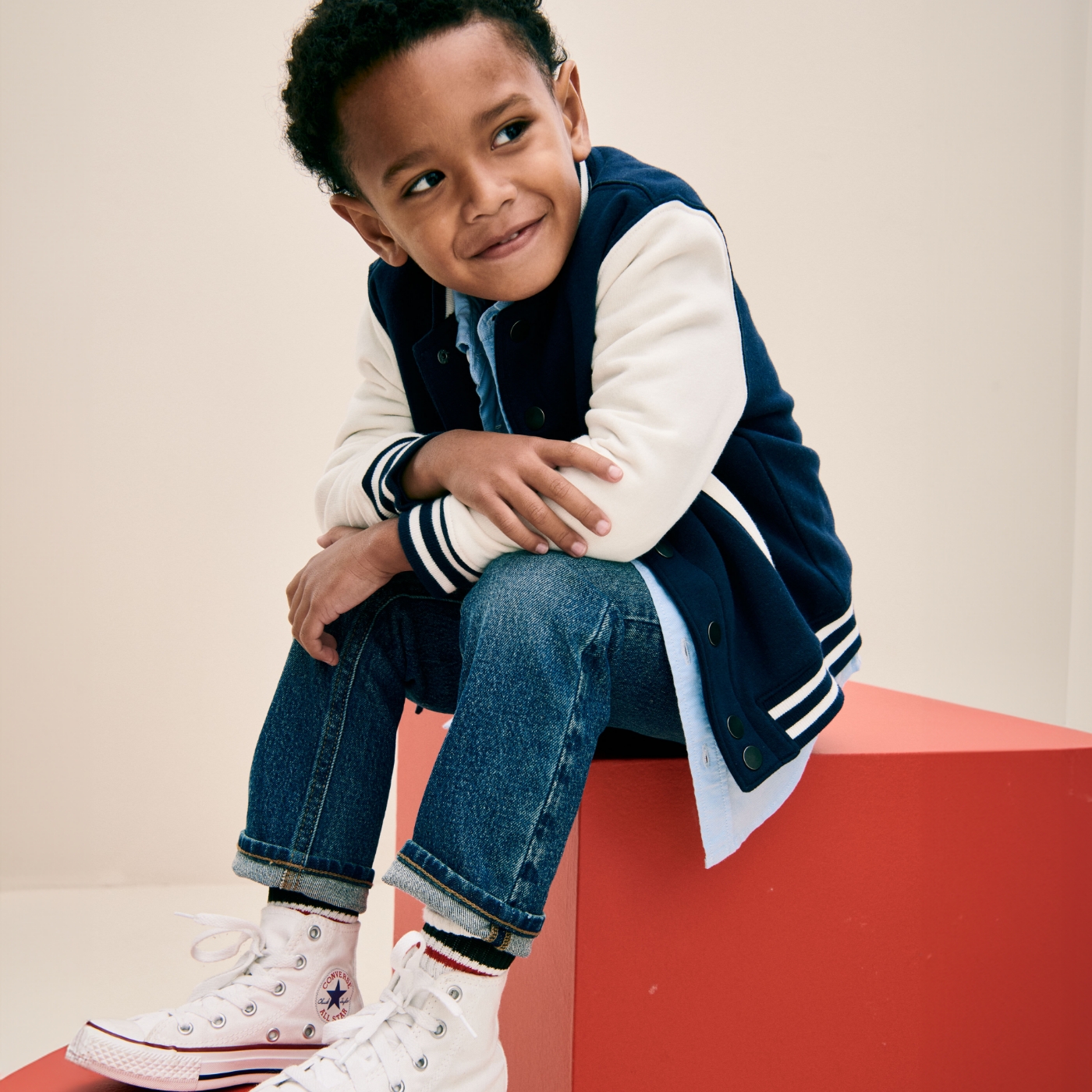 A toddler boy dressed in jeans, a letterman jacket, and hightop sneakers.