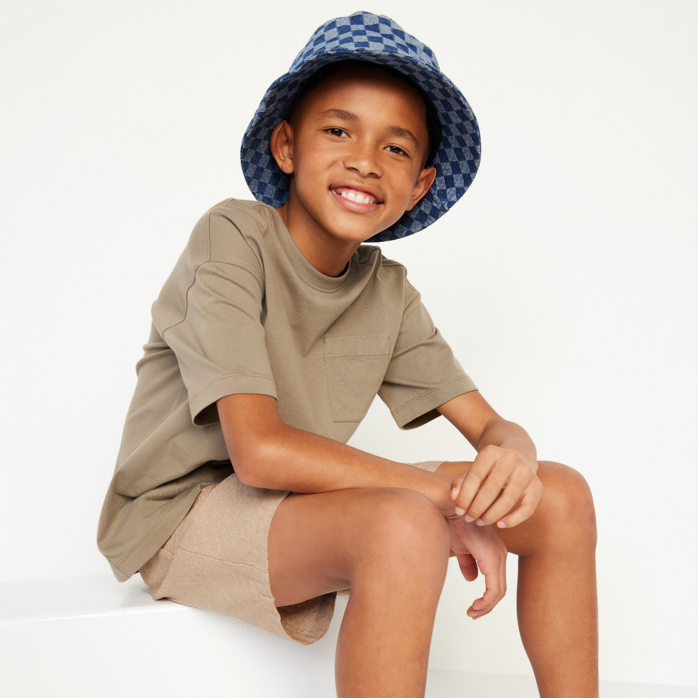 Young boy model wearing tan t-shirt and tan shorts with checkered bucket hat.