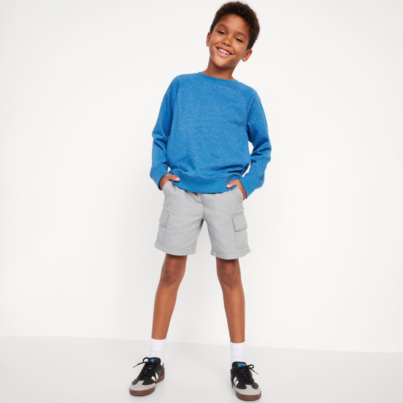 Un jeune garçon porte un t-shirt bleu ras du cou à manches longues et un short cargo gris.