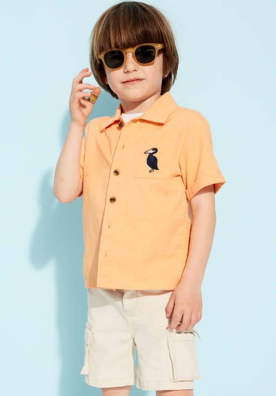Un jeune garçon porte une chemise orange et un short kaki cargo.